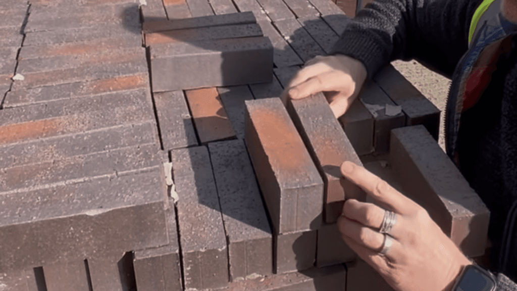Hands with rings examine two dark brown bricks pulled from a large, organized stack.