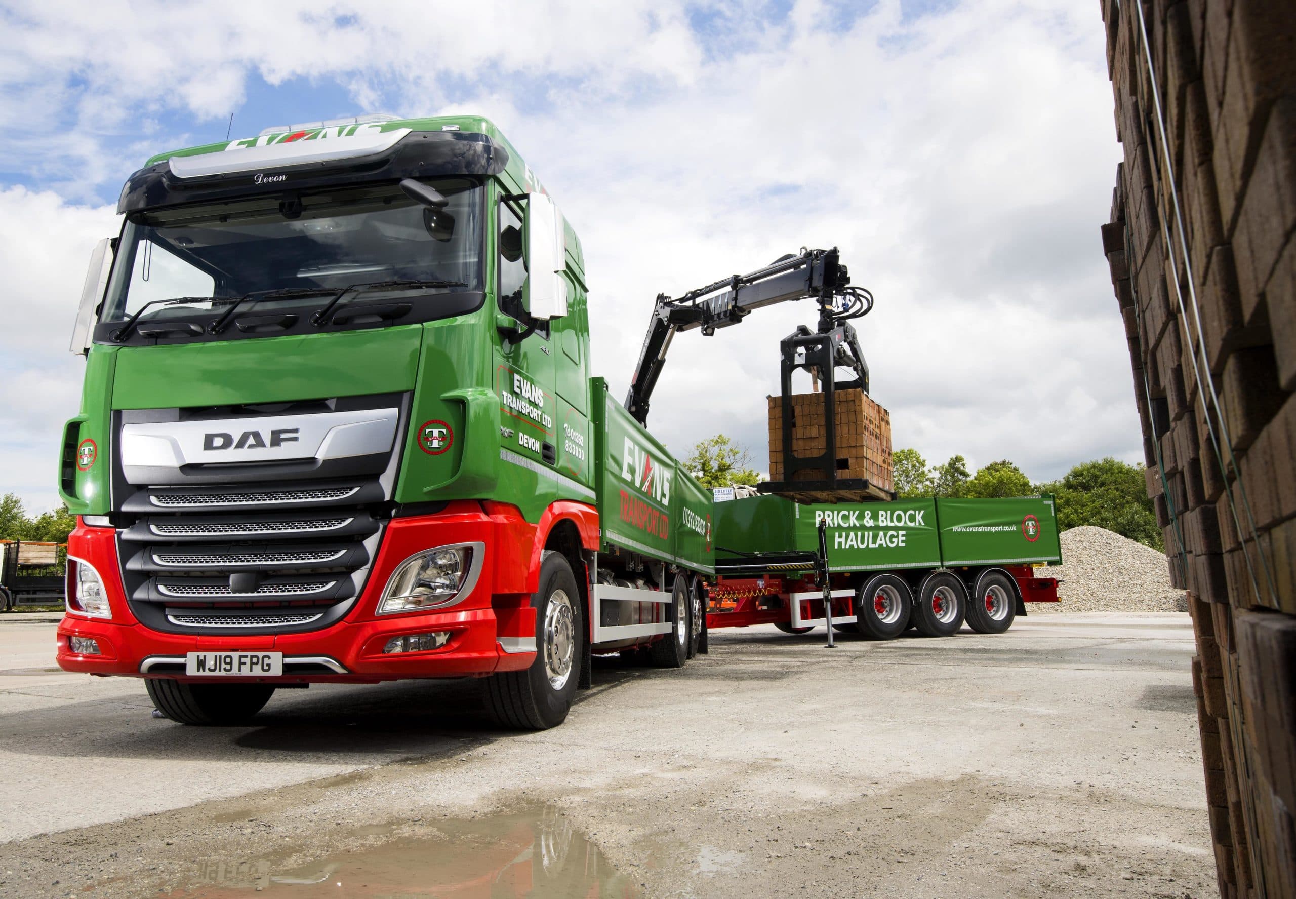 Green and red DAF truck using a crane to lift a pallet of bricks.