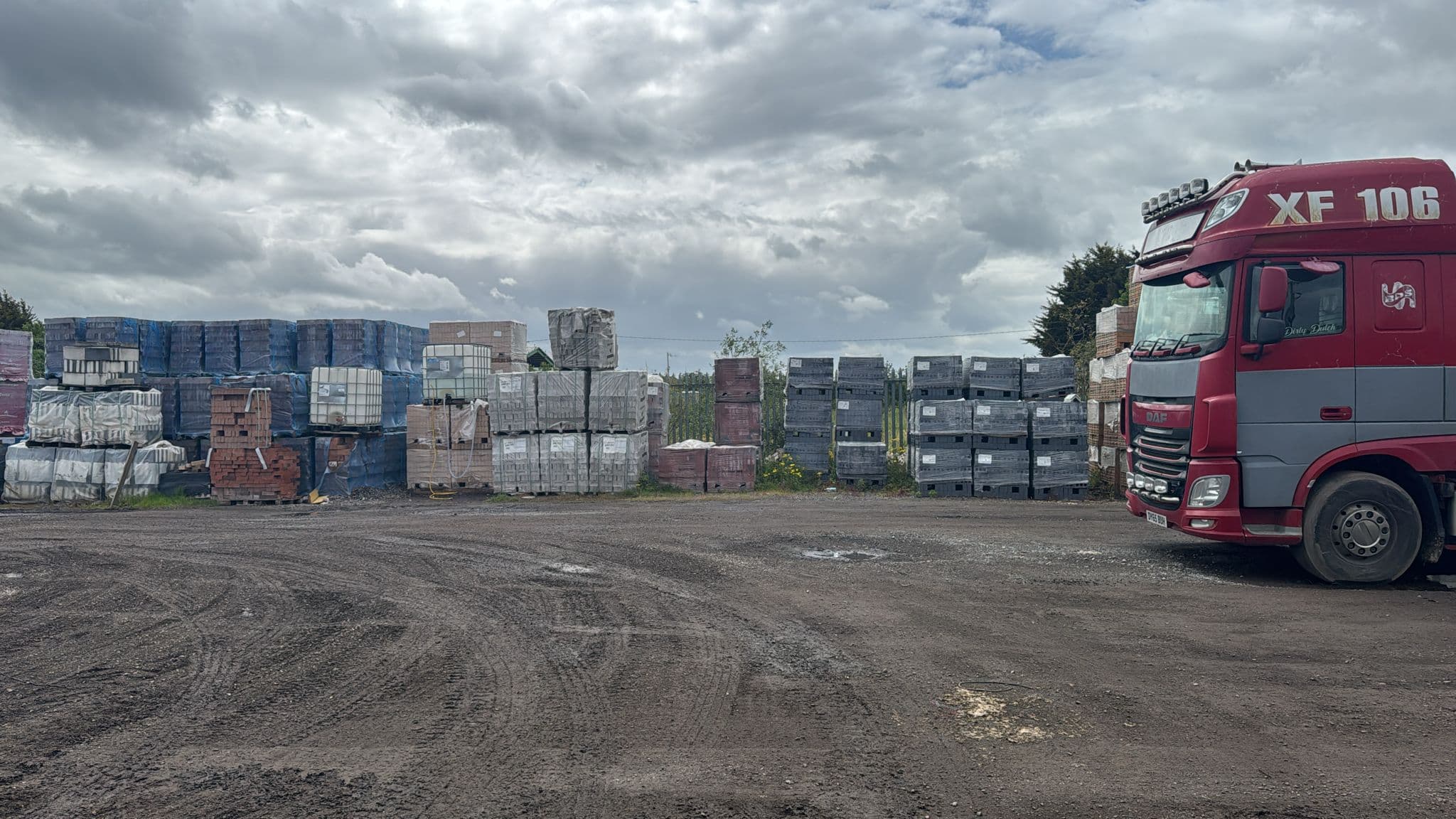 Red semi-truck parked in a yard beside stacks of building materials under a cloudy sky.