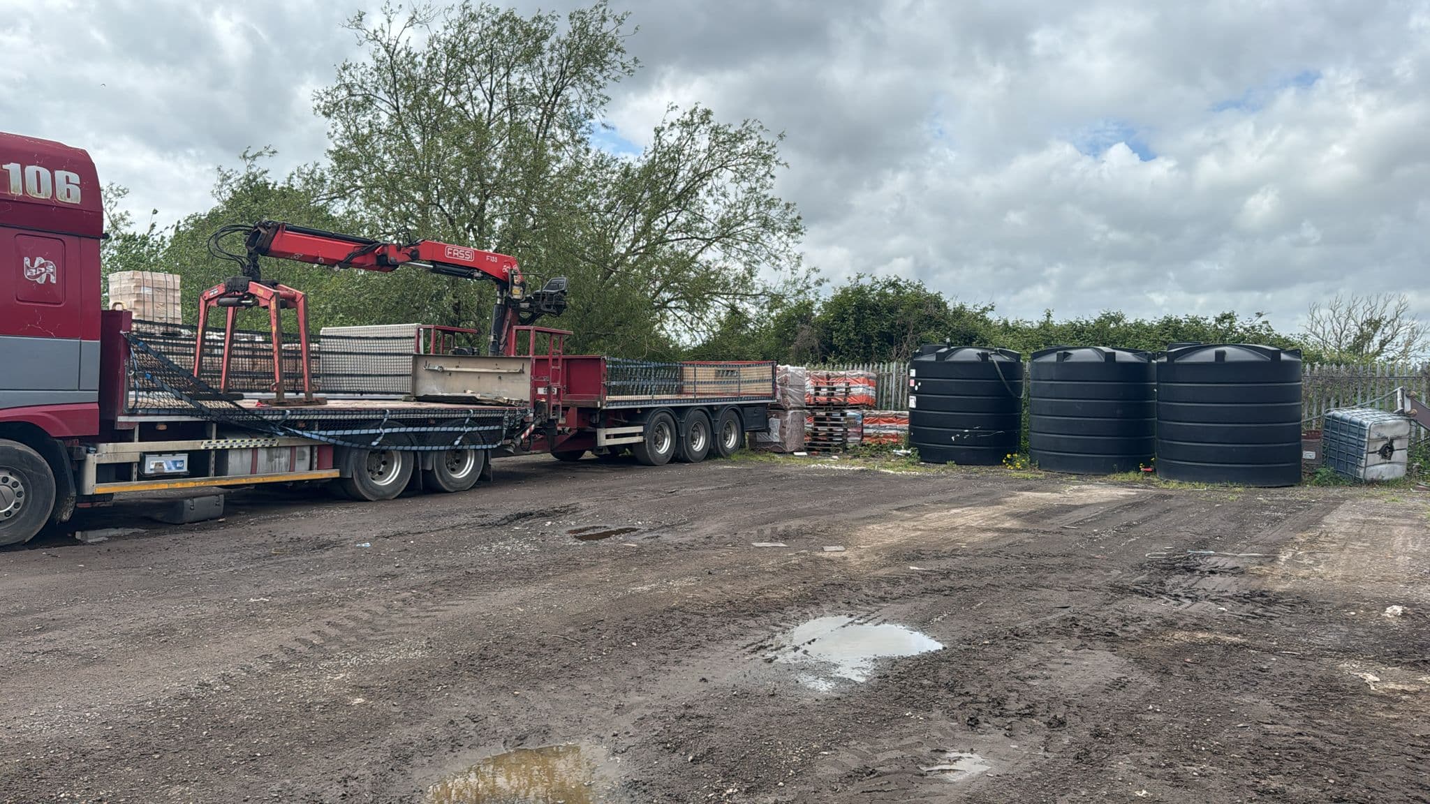 Red crane truck parked in a muddy yard next to three large black water tanks.
