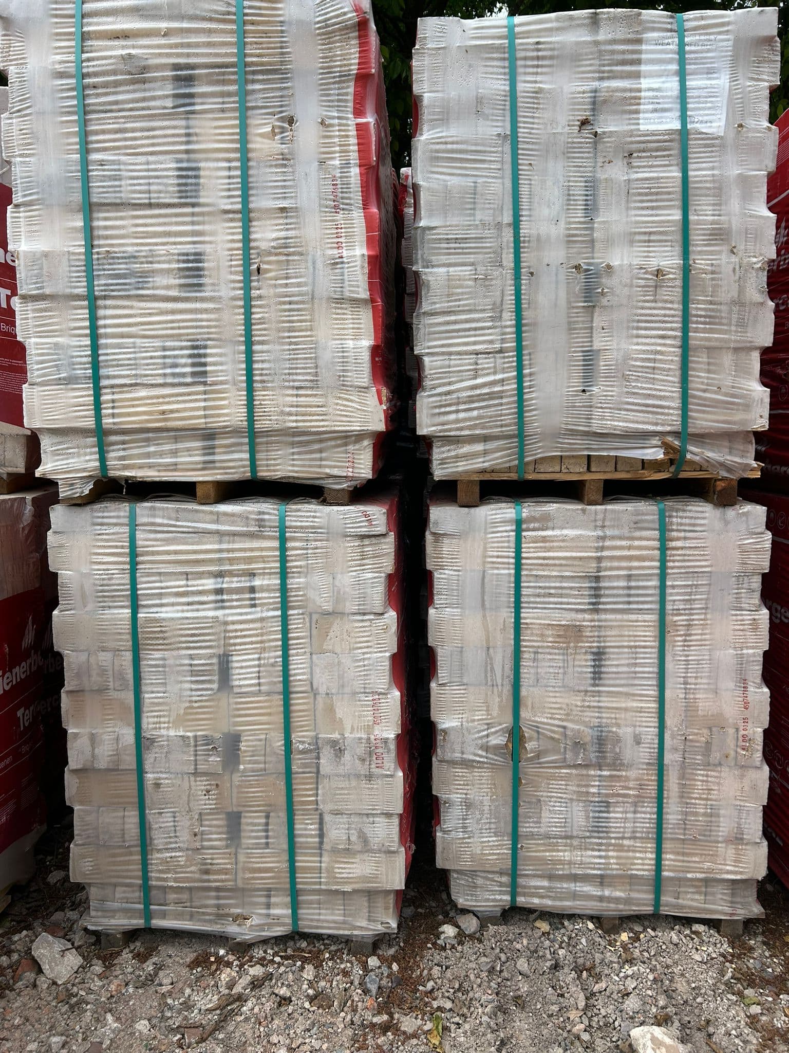 Four pallets of bricks wrapped in clear plastic and secured with green straps on gravel.